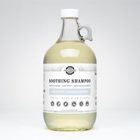 A glass jug displays Rustic Strength Soothing Shampoo, highlighting its unscented, hypoallergenic, and plant-derived formula.