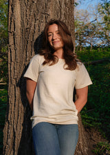 Woman smiles while leaning against a tree wearing a cream t-shirt and jeans in the woods.
