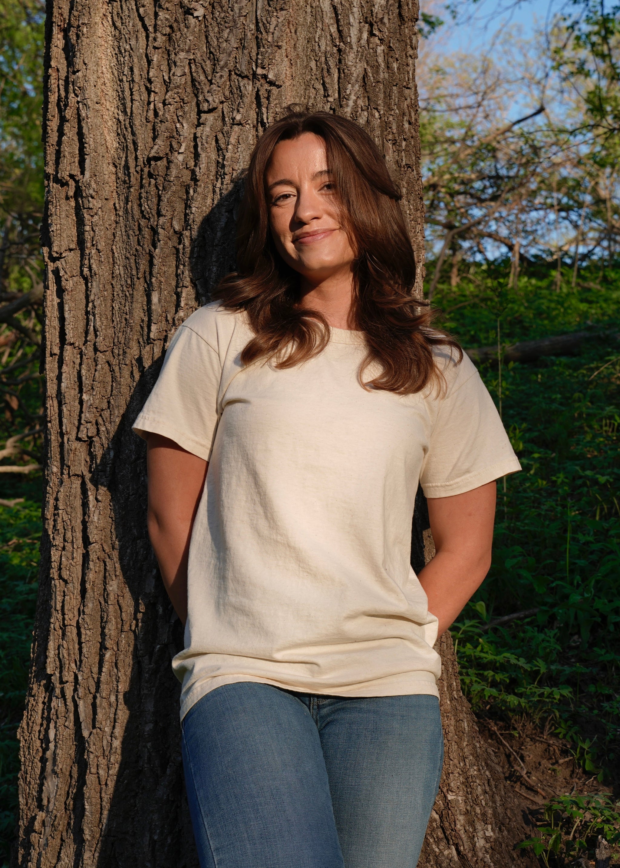 Woman smiles while leaning against a tree wearing a cream t-shirt and jeans in the woods.
