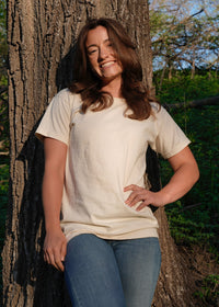 A smiling woman in a cream-colored t-shirt and jeans poses naturally against a large tree in the forest.

