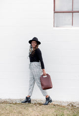 A stylish woman with long braids walks alongside a white wall, carrying a modern leather tote bag and wearing plaid trousers and a black hat.
