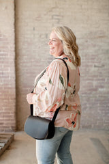 Smiling woman models a black leather crossbody bag and floral print blouse in front of a brick wall.
