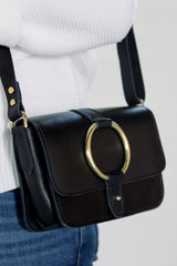 Person wears a black leather crossbody bag with a gold ring detail and a white sweater.
