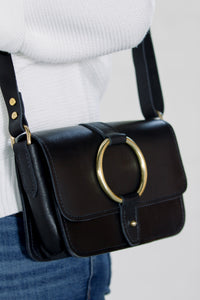 Person wears a black leather crossbody bag with a gold ring detail and a white sweater.
