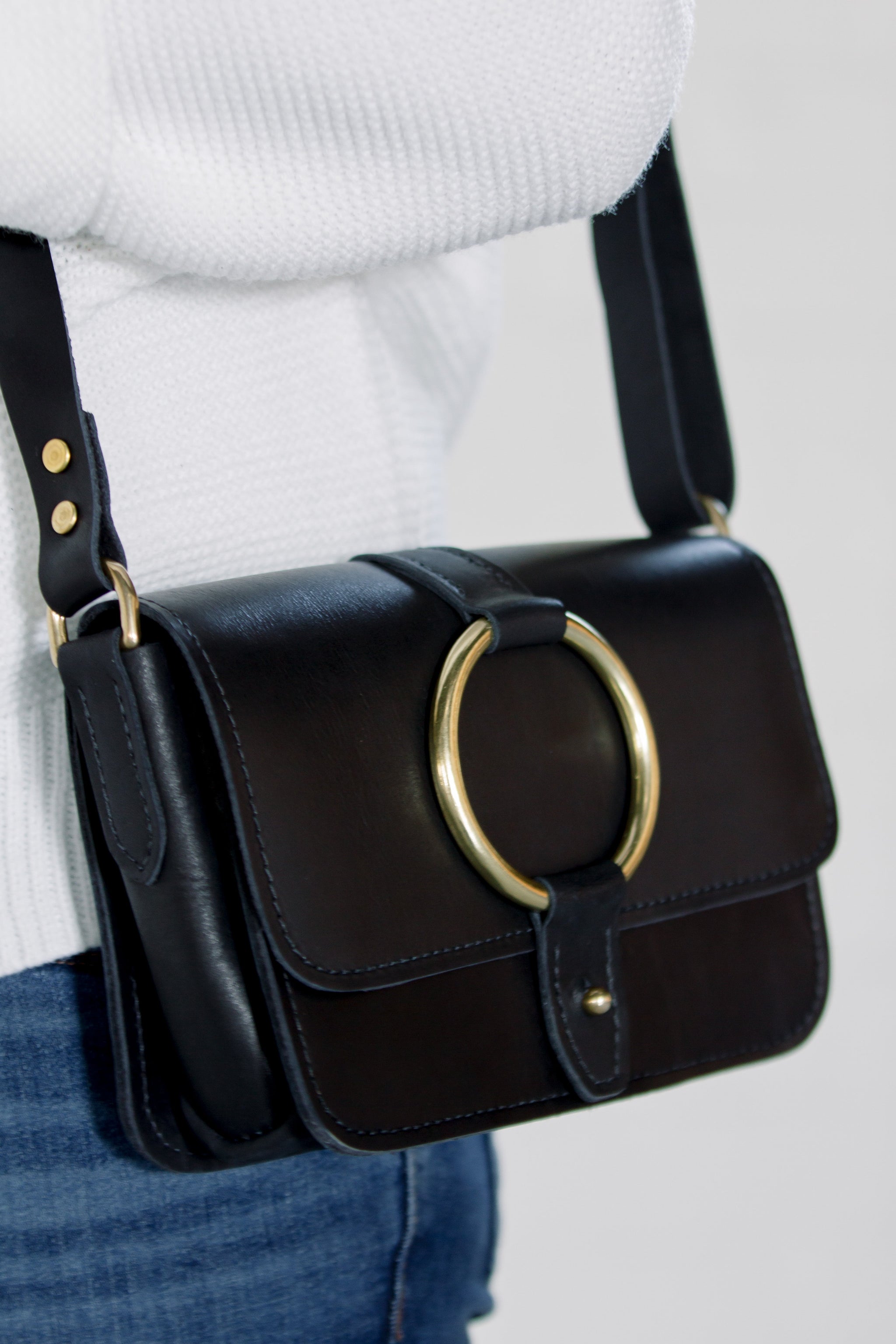 Person wears a black leather crossbody bag with a gold ring detail and a white sweater.
