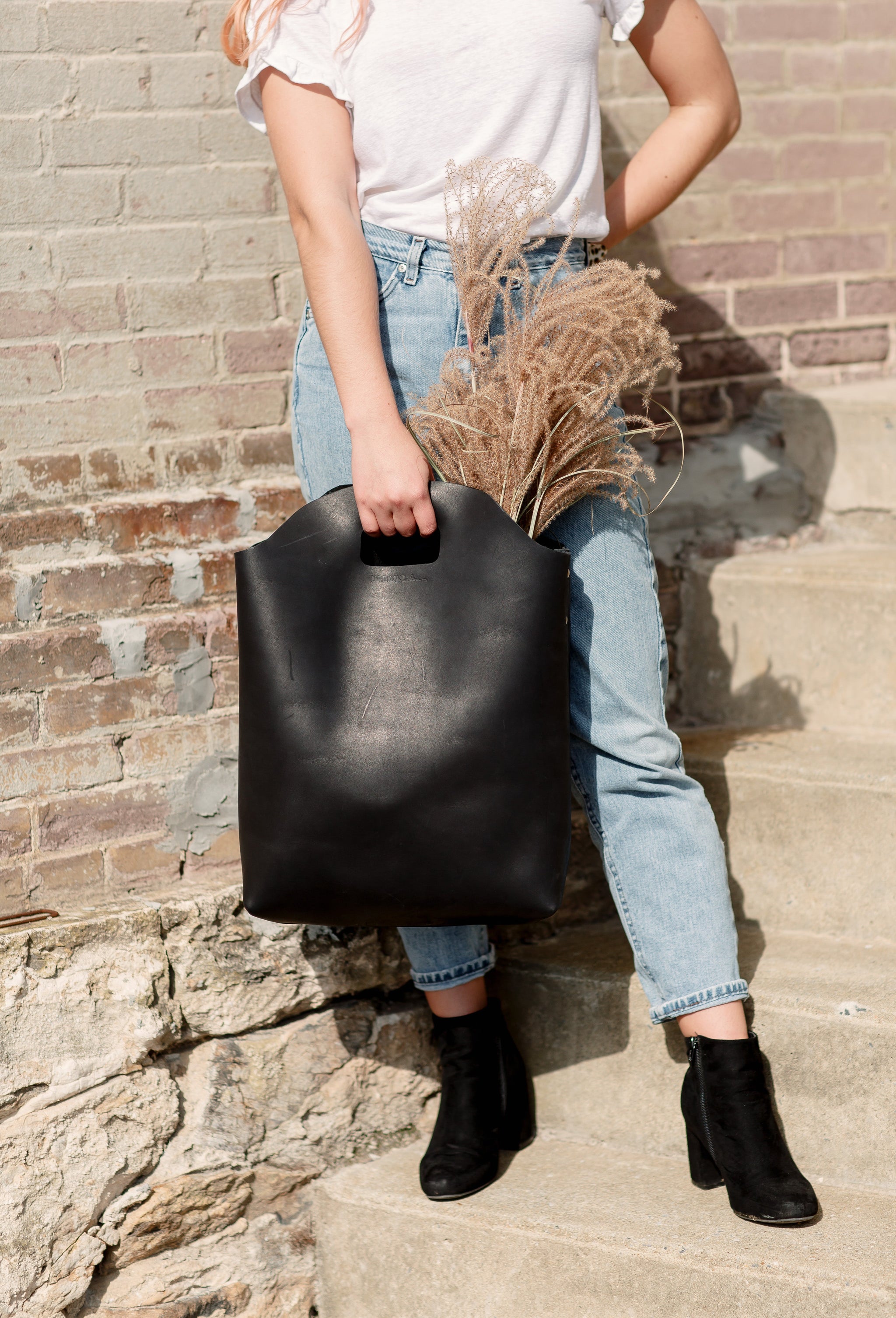 A woman carries a large black leather tote bag filled with pampas grass while standing on stone steps.
