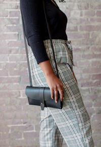 Woman showcases a black leather crossbody bag with a strap while wearing plaid paperbag waist pants and silver rings.
