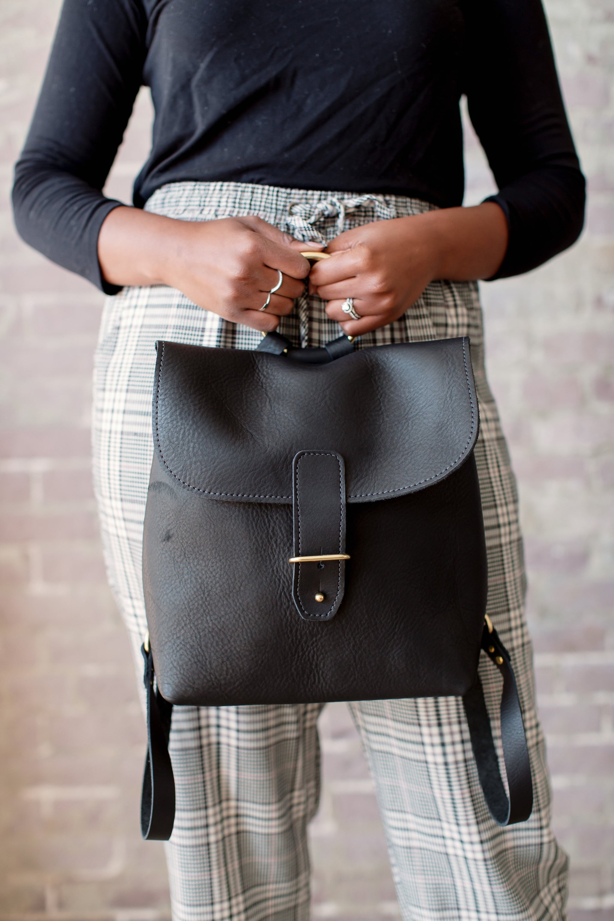 Woman showcases a stylish black leather backpack against a brick wall, wearing plaid pants.
