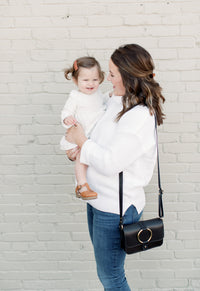 A stylish mother smiles while holding her happy toddler daughter in front of a white brick wall.
