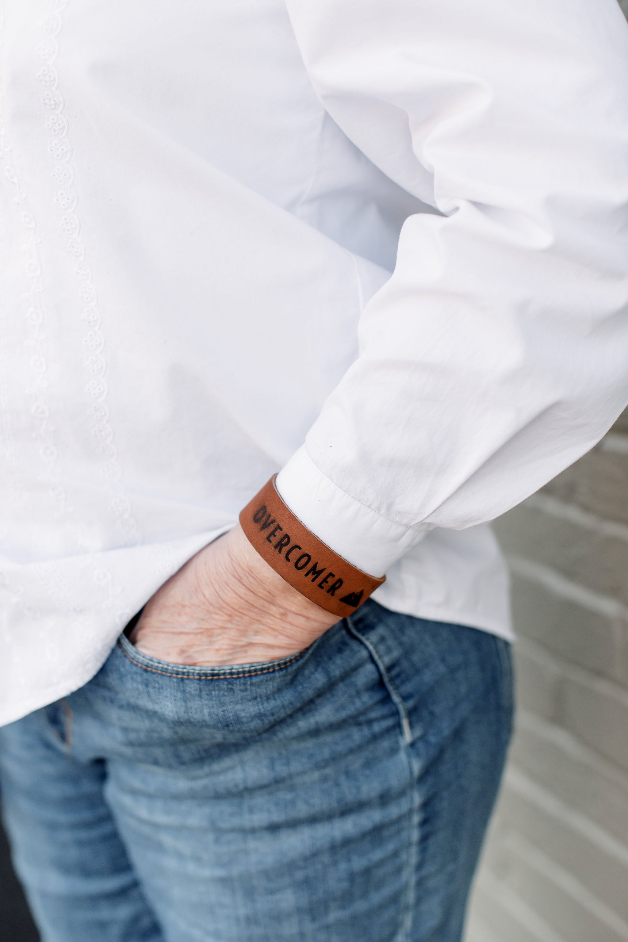 Person wears an "Overcomer" leather bracelet while putting a hand in their denim jeans pocket.
