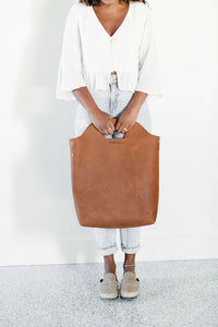 Woman models a brown leather Urban Southern tote bag, wearing a white top and light wash jeans.
