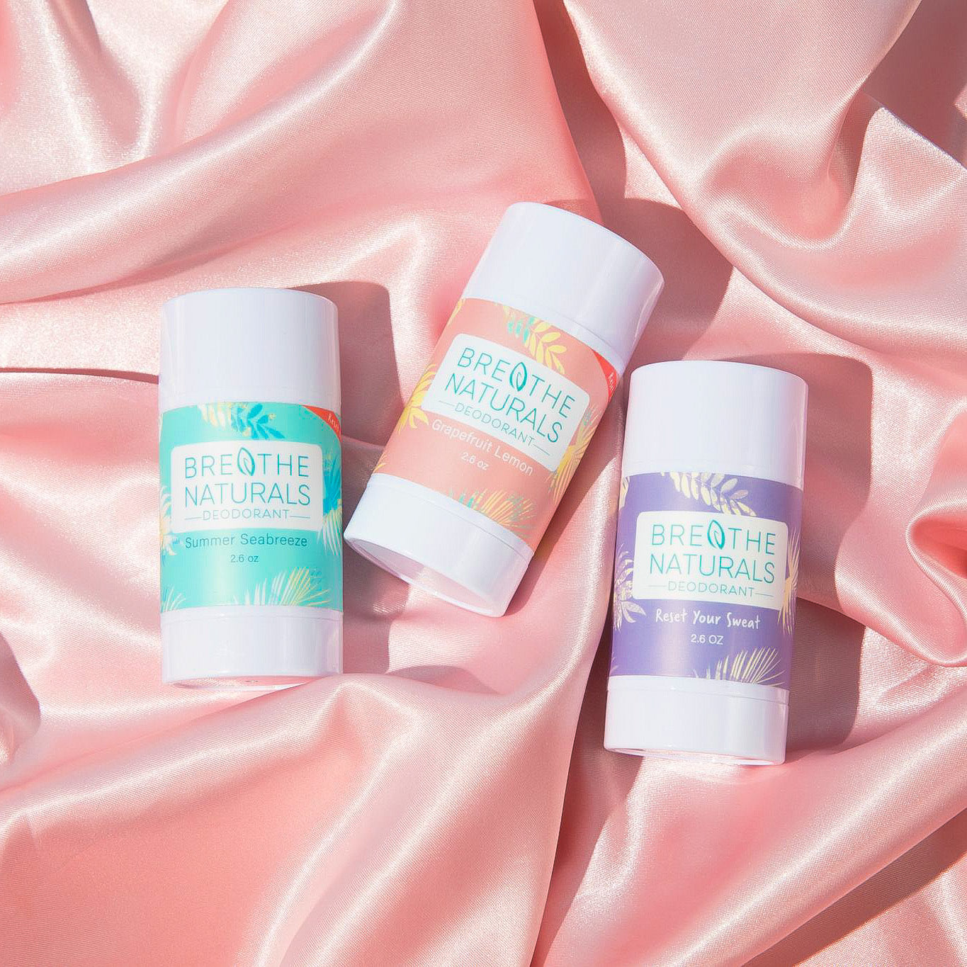 Breathe Naturals deodorant sticks displaying Summer Seabreeze, Grapefruit Lemon, and Reset Your Sweat scents sit on a pink satin fabric.
