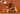 Three lit candles in amber glass jars from Fortana are arranged on wooden blocks against a warm brown background. A hand holds a small white pumpkin above the products, with additional pumpkins, an orange, cinnamon sticks, and vanilla beans styled around them.