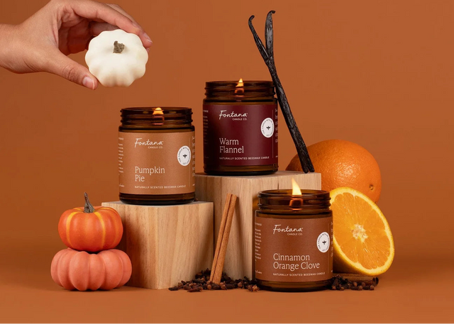 Three lit candles in amber glass jars from Fortana are arranged on wooden blocks against a warm brown background. A hand holds a small white pumpkin above the products, with additional pumpkins, an orange, cinnamon sticks, and vanilla beans styled around them.