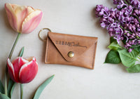 Urban Southern's handcrafted leather key pouch rests with colorful tulips and lilacs, showcasing a stylish everyday carry accessory.
