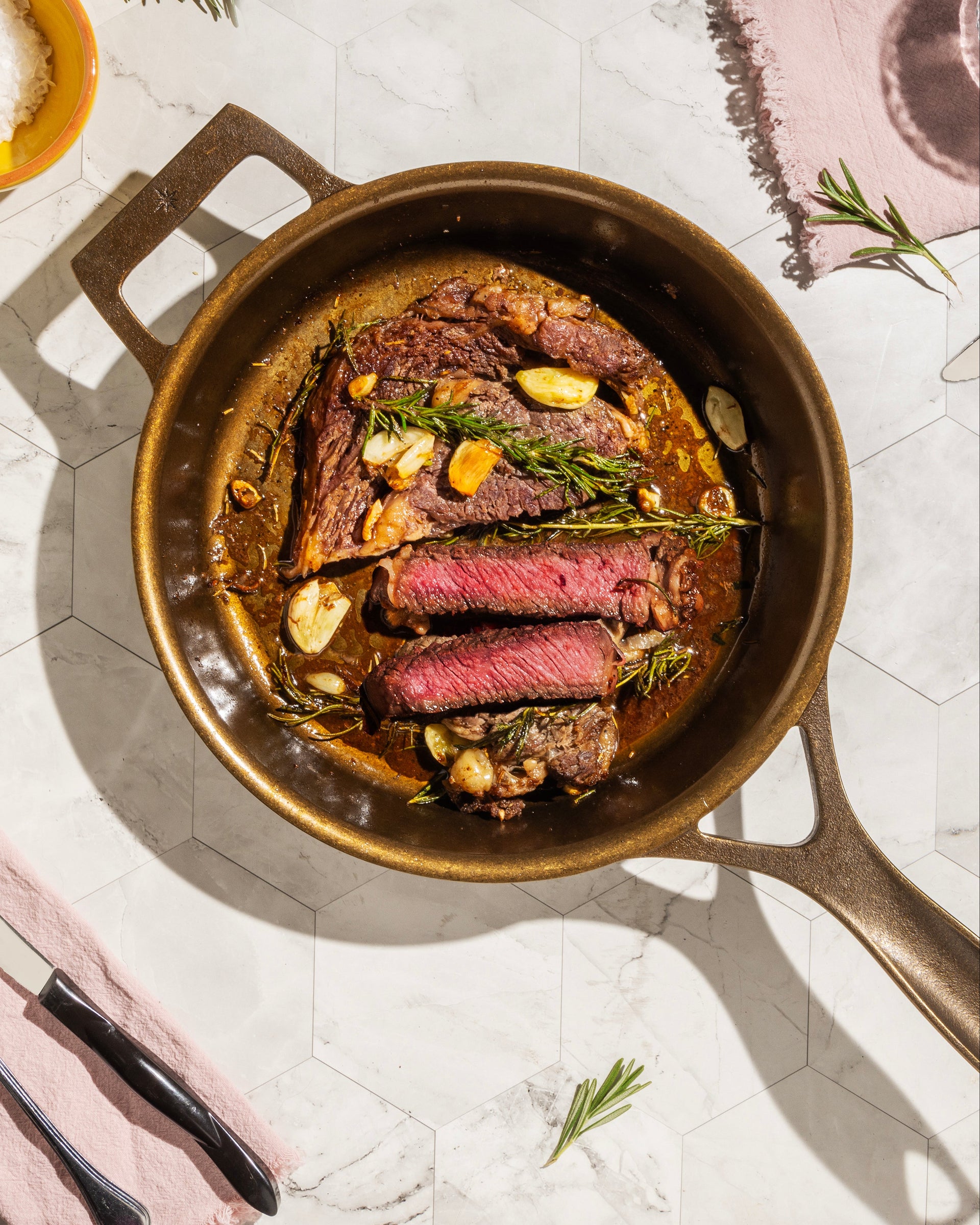 A top-down styled food scene showing cooked sliced steak in a bronze pan on a light marble surface, with plates of sliced steak, forks, knives, glasses of water, rosemary sprigs, and a small bowl of white salt around it.