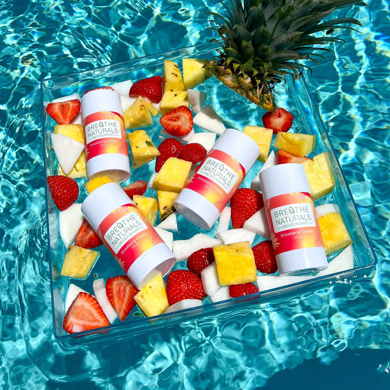 Breathe Naturals Strawberry Colada deodorant sticks float amongst pineapple, coconut, and strawberries in a clear tray on shimmering blue pool water.
