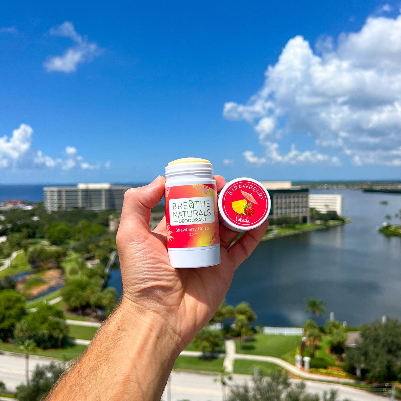 A hand holds Breathe Naturals Strawberry Colada deodorant against a scenic landscape with water and buildings.
