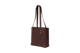 A studio shot presents a stylish, dark brown leather tote bag with brass rivet details and long shoulder straps.
