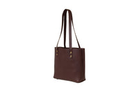 A studio shot presents a stylish, dark brown leather tote bag with brass rivet details and long shoulder straps.
