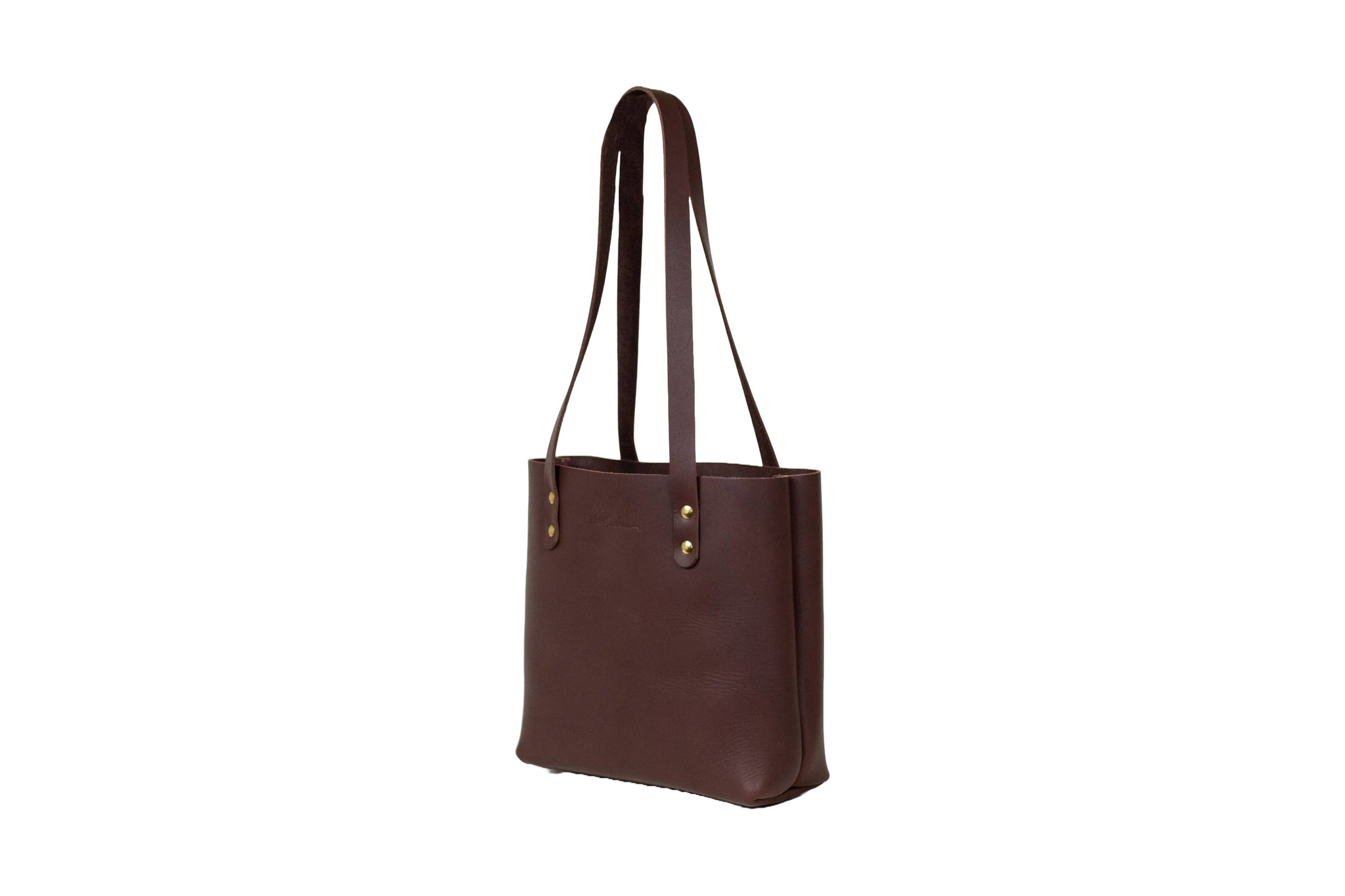 A studio shot presents a stylish, dark brown leather tote bag with brass rivet details and long shoulder straps.
