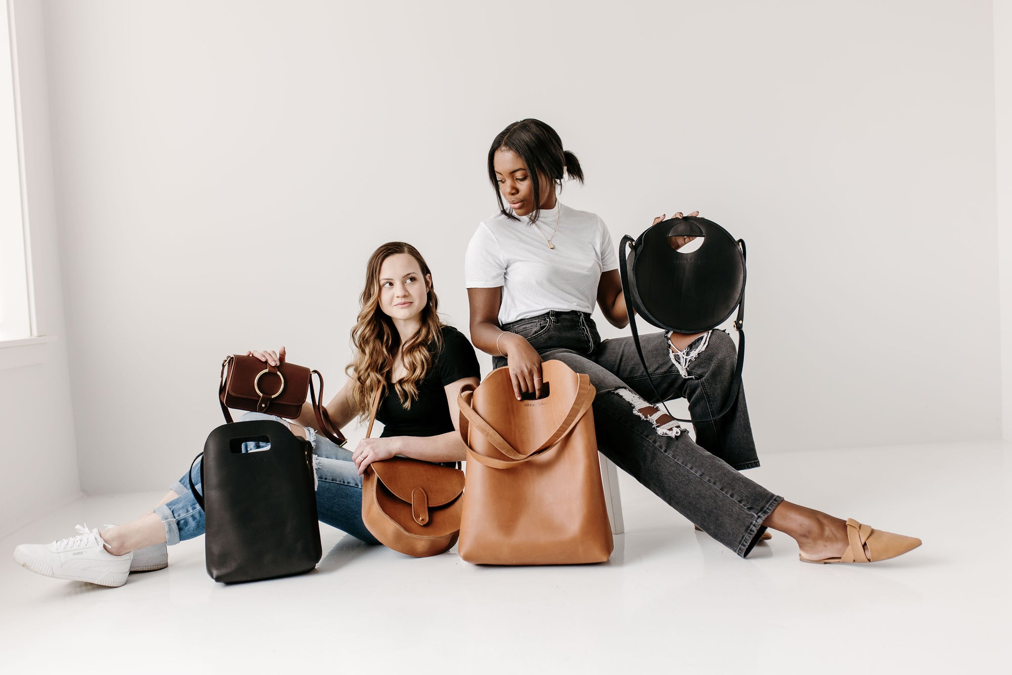 Two women display stylish leather handbags in a bright, minimalist studio setting.
