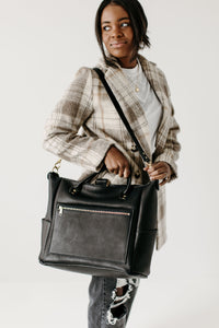 A stylish Black woman carries a black leather tote bag with a shoulder strap while wearing a plaid coat and ripped jeans.
