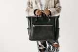 Woman models a stylish black leather tote bag with a front zipper pocket and side pockets.
