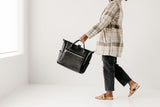 Woman carries a stylish black leather tote bag while walking in a plaid coat and distressed jeans.
