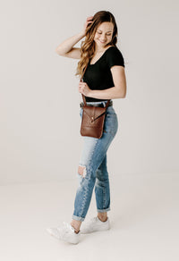 Woman smiles and adjusts her hair while modeling a brown leather crossbody bag with ripped jeans and white sneakers.
