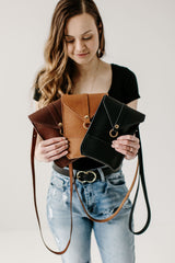 A woman displays three handmade leather crossbody bags in black, tan, and brown.
