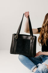 Woman displays a stylish black leather tote bag with gold buckle details.
