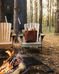 A leather tote bag filled with firewood sits on an Adirondack chair near a crackling campfire at a forest campsite.
