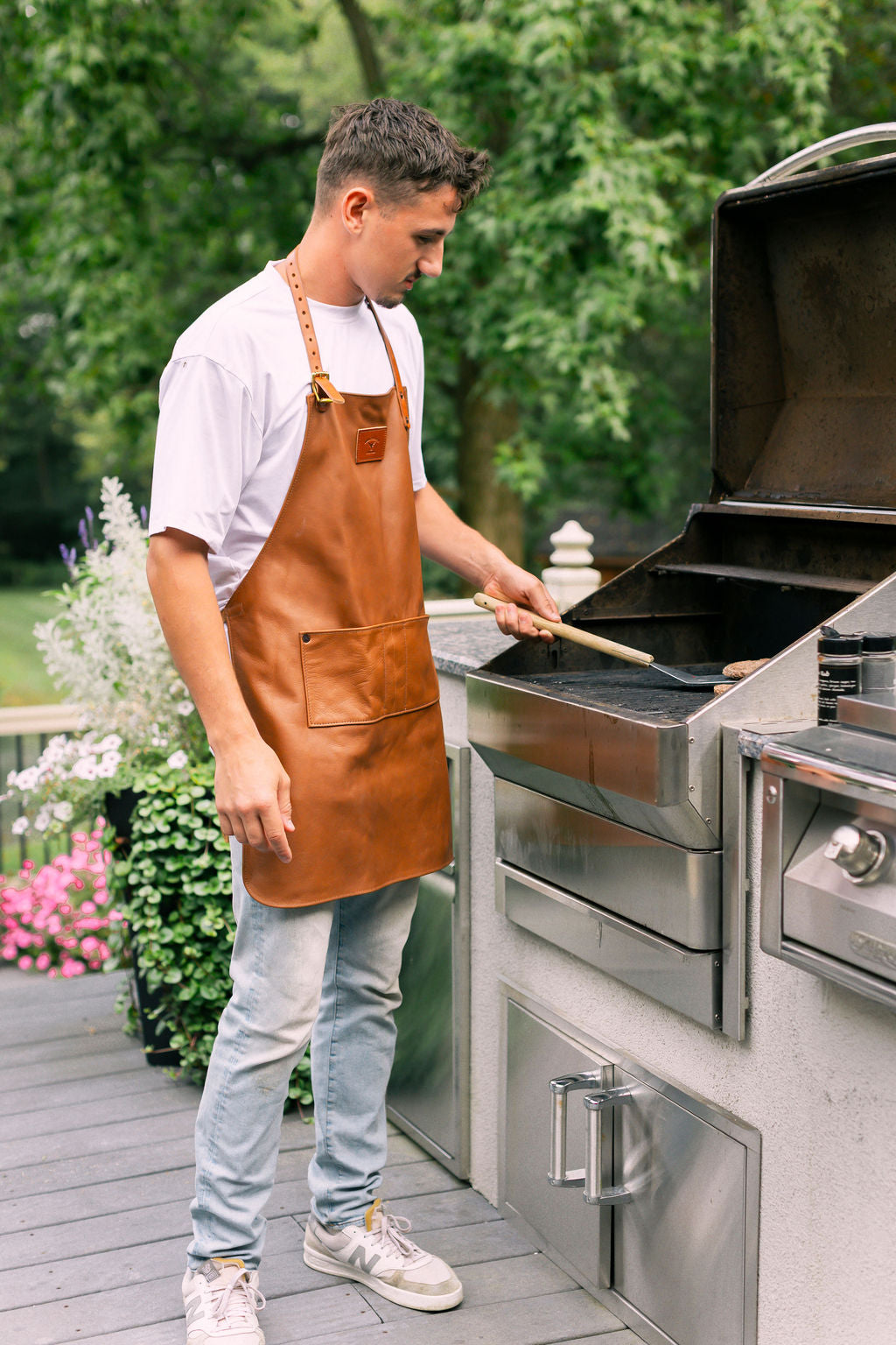 A man wearing a leather apron grills burgers outdoors on a stainless steel barbecue.
