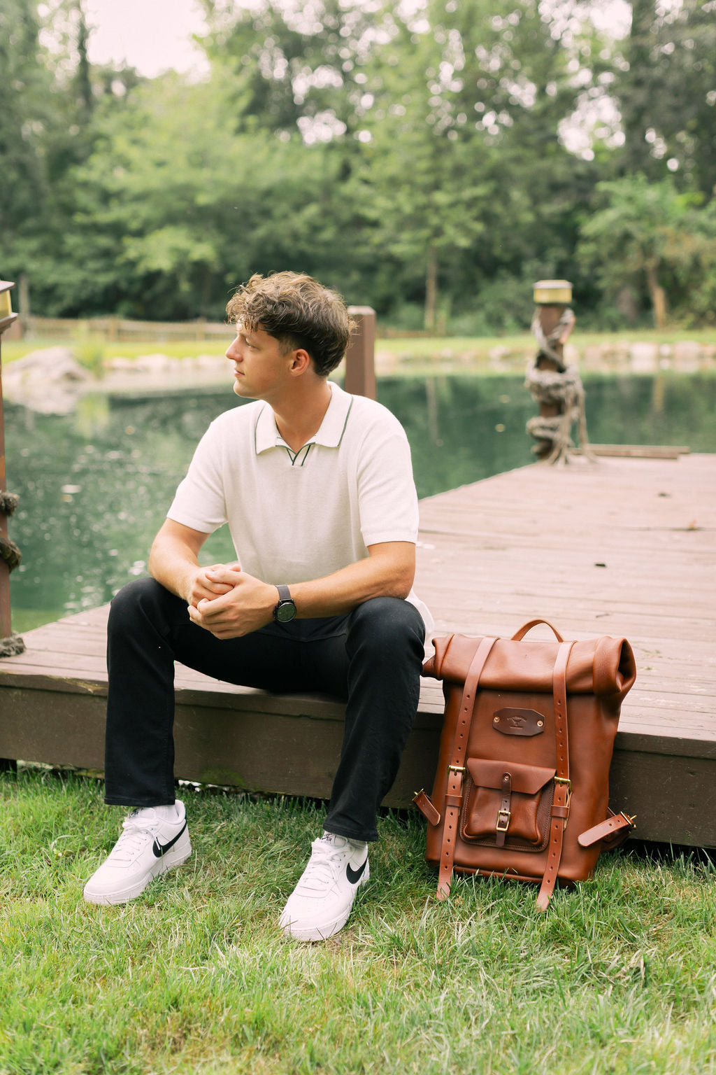 Man in a polo shirt sits on a wooden dock with a leather backpack and Nike Air Force 1 sneakers, gazing at the lake.
