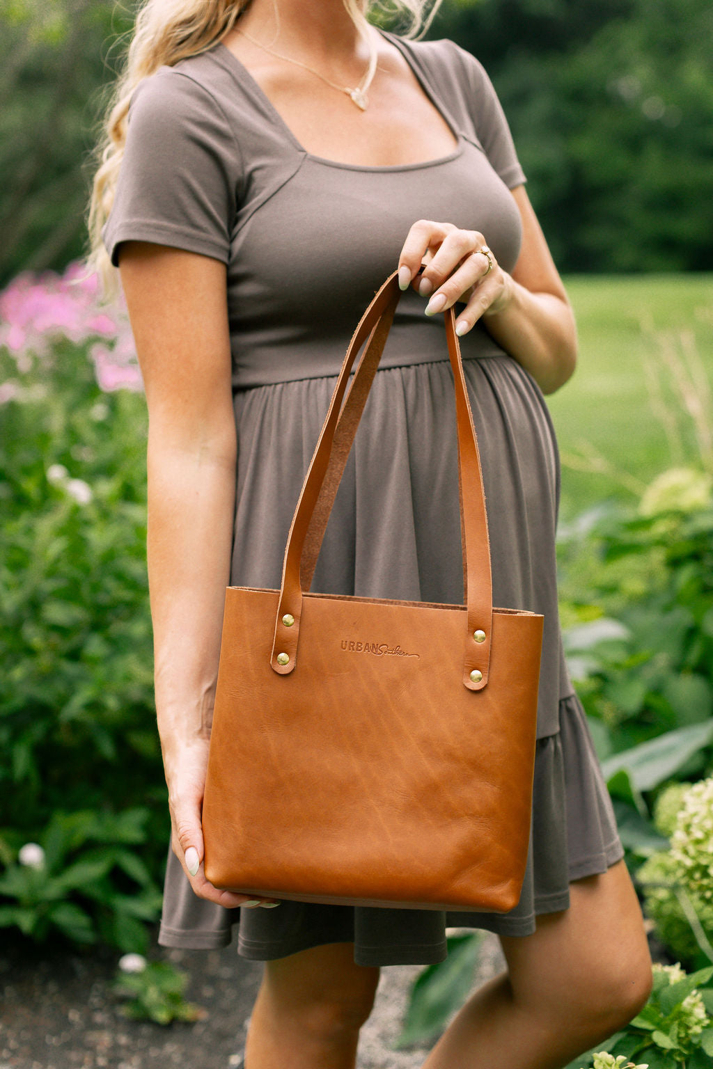 A pregnant woman showcases an Urban Leather tote bag in an outdoor setting.
