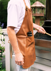 A man wearing a leather grilling apron and holding a spatula stands by a barbecue, seasoning in his pocket.
