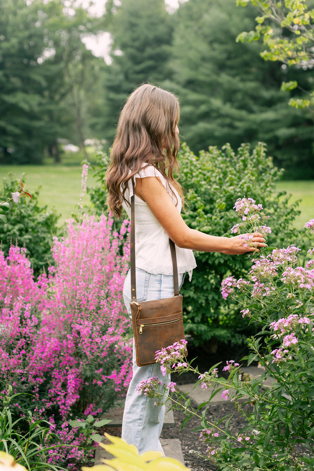 A woman wearing a leather crossbody bag gently touches pink flowers in a lush garden setting.
