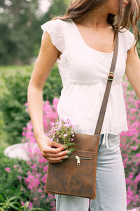 Woman adds delicate pink flowers to her brown leather crossbody bag in a lush garden setting.
