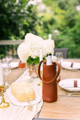 Champagne stays chilled in a leather wine cooler on an elegant outdoor dining table setting with white hydrangeas and fresh bread.
