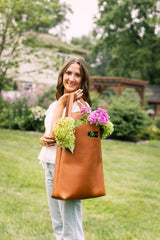 A smiling woman carries a brown leather tote bag overflowing with fresh hydrangea flowers in a garden setting.
