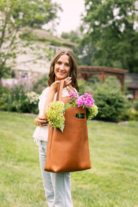 A smiling woman carries a brown leather tote bag overflowing with fresh hydrangea flowers in a garden setting.
