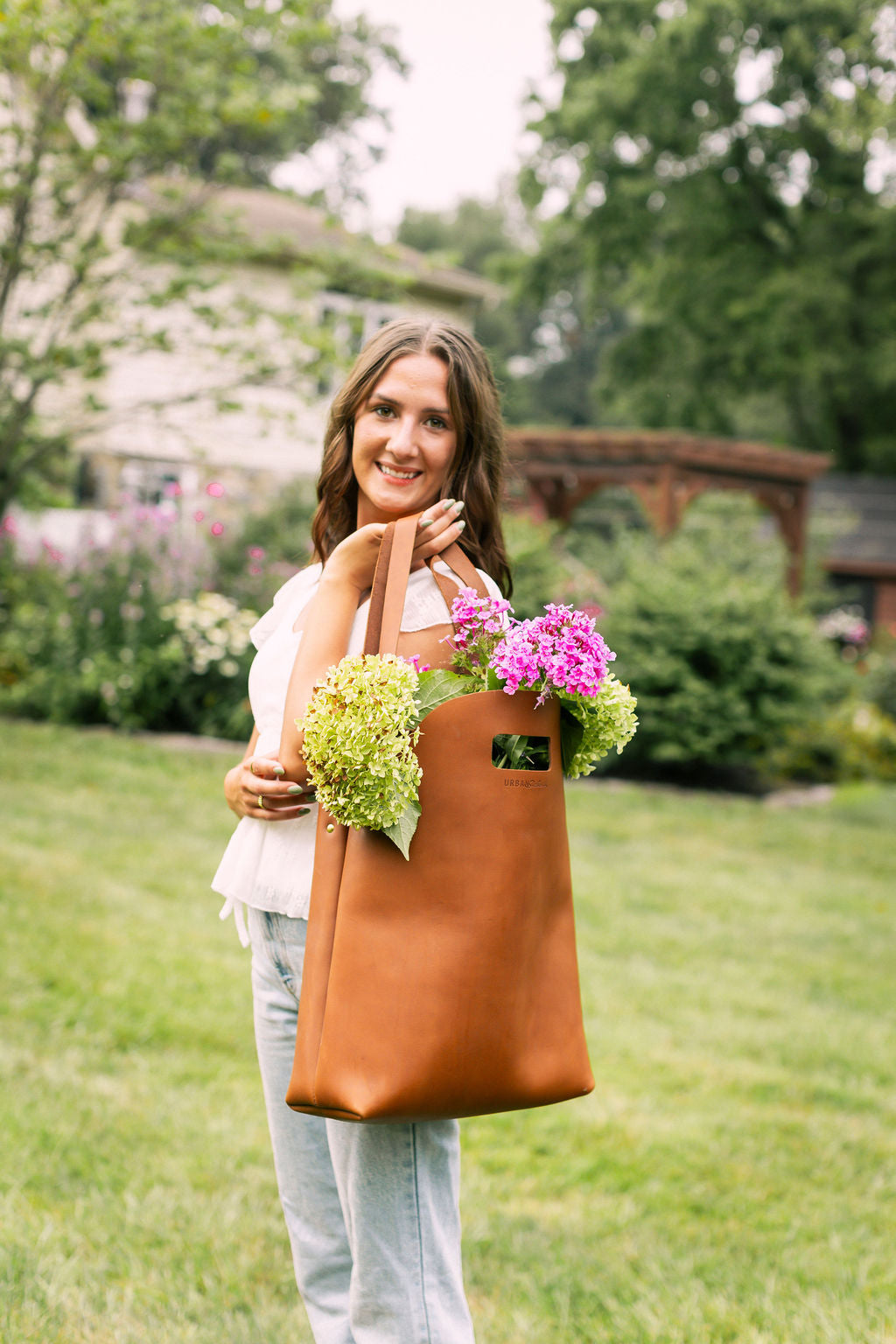 A smiling woman carries a brown leather tote bag overflowing with fresh hydrangea flowers in a garden setting.

