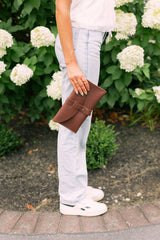 A woman models a brown leather Urban Skin clutch purse while standing near a hydrangea bush.
