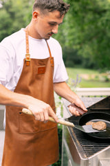 Man wearing a leather apron grills juicy hamburgers on an outdoor barbecue.
