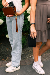 Two women showcase leather phone wallets with wristlets outside among greenery.
