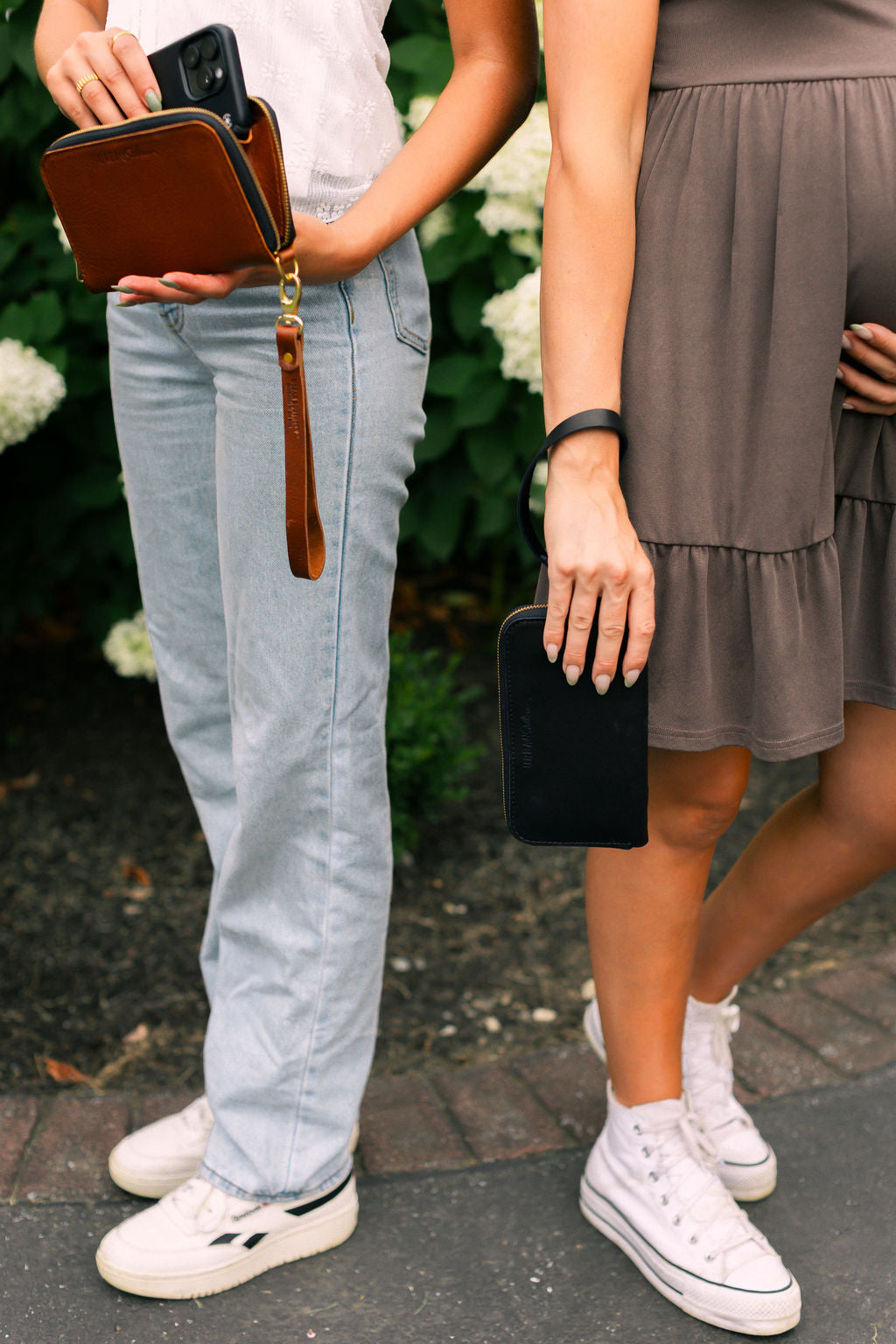 Two women showcase leather phone wallets with wristlets outside among greenery.
