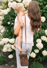 A mother carrying her toddler wears a leather crossbody bag in front of a white hydrangea bush.
