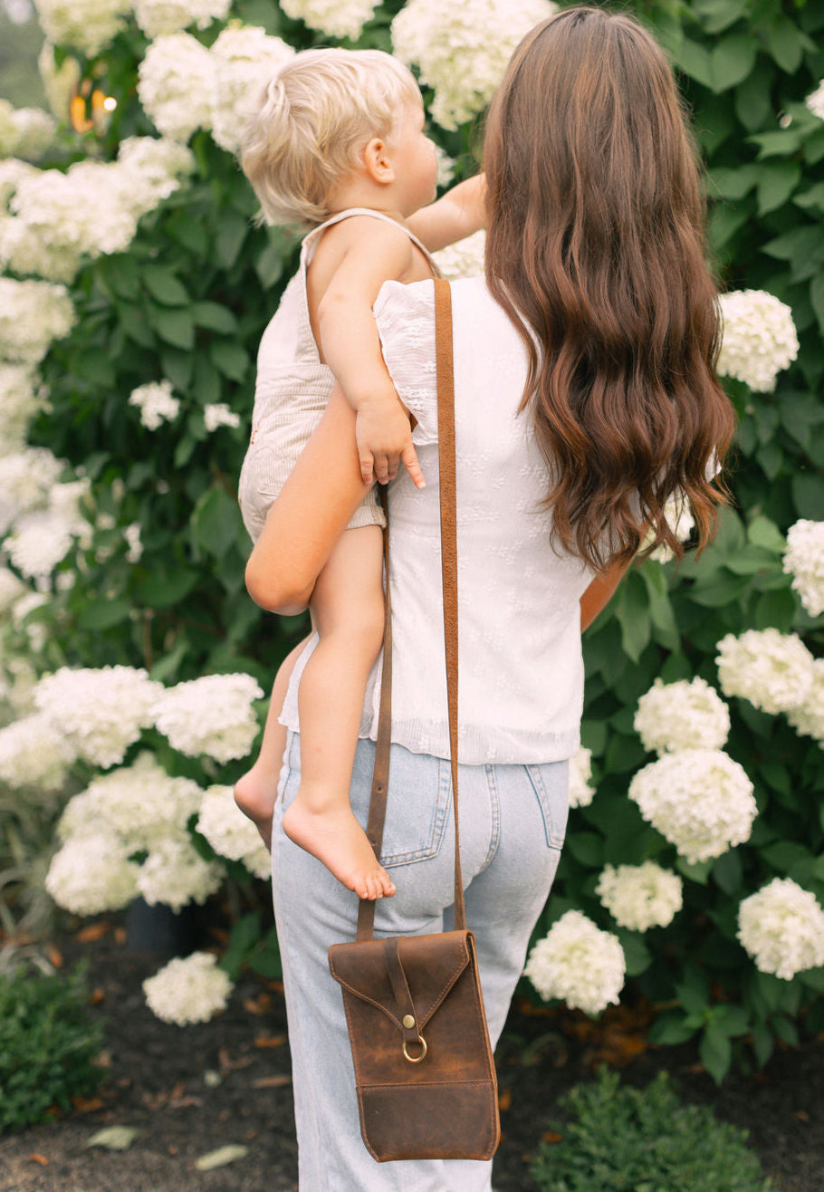 A mother carrying her toddler wears a leather crossbody bag in front of a white hydrangea bush.
