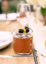 A refreshing blackberry cocktail, garnished with lemon, sits in a leather-sleeved glass on a stylish outdoor table setting.
